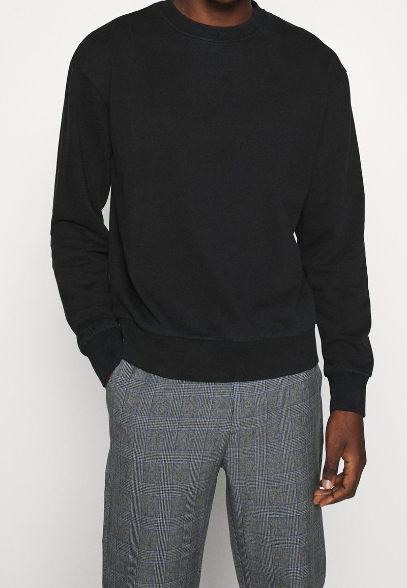 Sudadera negra con cuello redondo, mangas largas y puños acanalados, combinada con pantalones de cuadros grises con acentos azules.