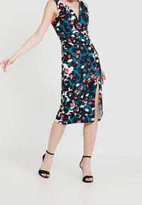 Robe portefeuille sans manches noire avec un motif léopard coloré comprenant du bleu, du rouge et du blanc. Fente latérale à l'ourlet, assortie à des talons noirs.