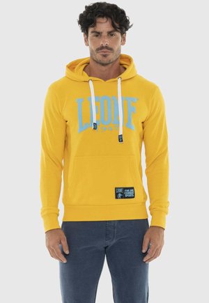 Uomo che indossa una felpa gialla con la scritta "LEONE 1947" e jeans blu, in piedi contro uno sfondo chiaro e semplice.