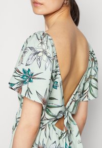 Vestido envelope floral em tecido verde claro, com mangas curtas bufantes, costas abertas com detalhe de laço e cintura ajustada.