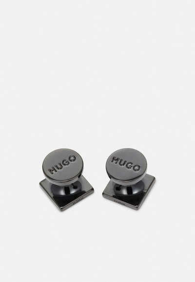 Boutons de manchette noirs ronds avec une finition brillante et le logo "HUGO" gravé, montés sur une base carrée. Le matériau semble métallique.