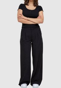 Černý zkrácený top s kulatým výstřihem a detailem knoflíků, kombinovaný s volnými, černými plisovanými kalhotami. Oba vyrobeny z jemného, hladkého materiálu.