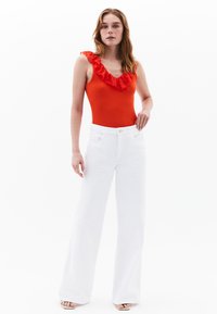 Body con scollo a V con rouches rosso abbinato a jeans bianchi a gamba larga. Materiale: tessuto morbido. Dettagli notevoli: rouches sul collo, bottone decorativo sui jeans.