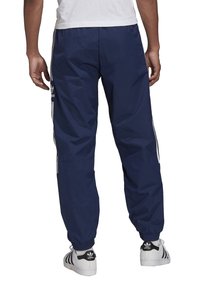 Námořnické modré sportovní kalhoty s bílými bočními pruhy, elastickými manžetami a lehkým materiálem, zobrazené zezadu s tričkem s kulatým výstřihem.