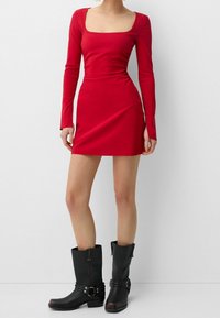 Rød langærmet kjole med firkantet halsudskæring, tætsiddende design og minilængde. Parret med sorte læder støvler til knæene med spændes detaljer.