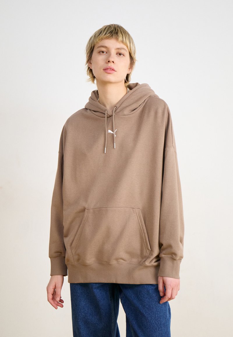 Felpa con cappuccio marrone, dotata di tasca frontale e polsini a costine. Presenta un piccolo logo bianco sul petto e una vestibilità morbida. Abbinata a jeans blu.