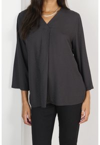 Černá blůza s krátkým rukávem a výstřihem do V, s jemnou texturou teček. Lehká tkanina s volným střihem, kombinovaná s černými kalhotami. Stříbrné šperky jako doplněk.