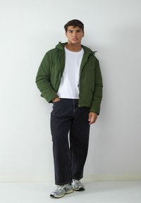 Giacca a vento verde con cappuccio, t-shirt bianca sotto, jeans in denim scuro e sneakers sportive argentate. Design semplice e funzionale.