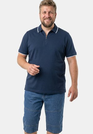 Hombre sonriente con cabello rubio corto que lleva una camisa polo azul marino de manga corta y pantalones cortos vaqueros azules hasta la rodilla, de pie, mirando hacia adelante.