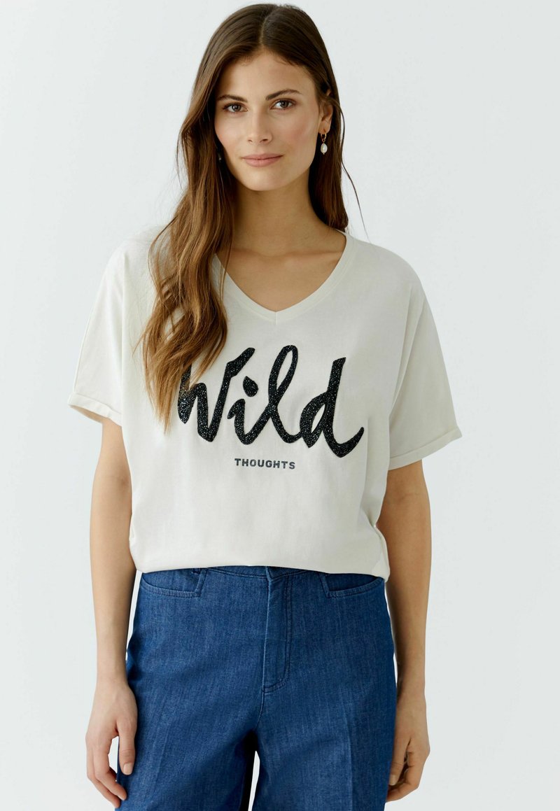 Helles beige V-Ausschnitt-T-Shirt mit "Wild" in schwarzem Glitzertext und "THOUGHTS" darunter in kleinerer Schrift. Kombiniert mit dunkelblauen Jeans.
