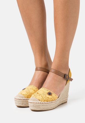 Sandales compensées en crochet jaune avec une bride de cheville marron, semelle en jute tissée naturelle et détail de bouton décoratif à l'avant.