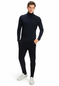 Marineblauer Rollkragenpullover mit Rippstruktur, kombiniert mit passenden schmal geschnittenen Hosen und weißen Sneakers, vor einem einfarbigen Hintergrund stehend.