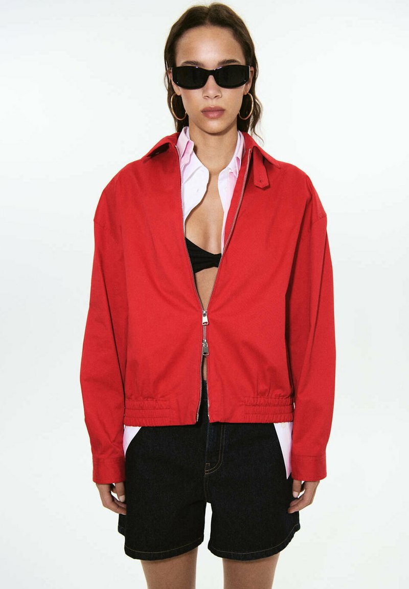Femme portant des lunettes de soleil noires, des boucles d'oreilles créoles dorées, une veste rouge zippée sur une chemise blanche et un soutien-gorge noir, assortie à un short noir.
