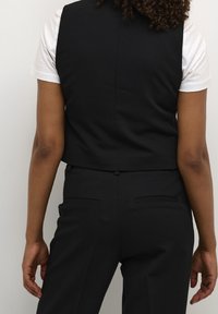 Gilet nero corto sopra una camicia bianca, caratterizzato da un design strutturato con schiena senza cuciture e tasche sottili sui pantaloni su misura.