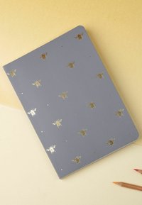 Šedý notebook se zlatými vzory včel a malými zlatými tečkami, s hladkým víkem a zaoblenými rohy. Příbor jsou částečně viditelné poblíž.