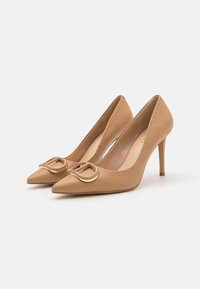 Beige ādas augstpapēžu kurpes ar norādītu purngalu, plānu stiletto papēdi un zelta sprādzi priekšpusē. Gluda tekstūra un minimālisma dizains.