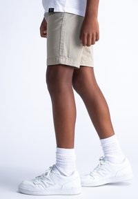 Pantaloni corti beige con orlo risvoltato, indossati con calze bianche a crew e sneakers bianche, caratterizzati da un'etichetta con logo sull'orlo della maglietta.