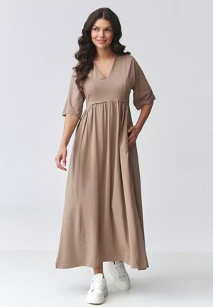 Kobieta o długich ciemnych włosach, ubrana w beżową sukienkę maxi z dekoltem w serek i białe trampki, stojąca na tle jednolitego jasnego tła.