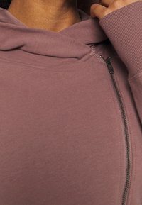 Zip-up hættetrøje lavet af blød, struktureret stof i en dæmpet mauve farve. Har en kengurulomme og ribbet manchetter. Lynlåslukning er synlig.