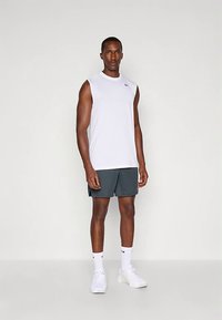 Witte mouwloze sportshirt, grijze shorts, witte sneakers en witte sokken met zwarte accenten. Model staat tegen een neutrale achtergrond.