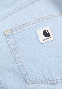 Tissu en denim bleu clair avec une poche arrière visible et des coutures orange, arborant une étiquette de logo Carhartt noir et blanc.