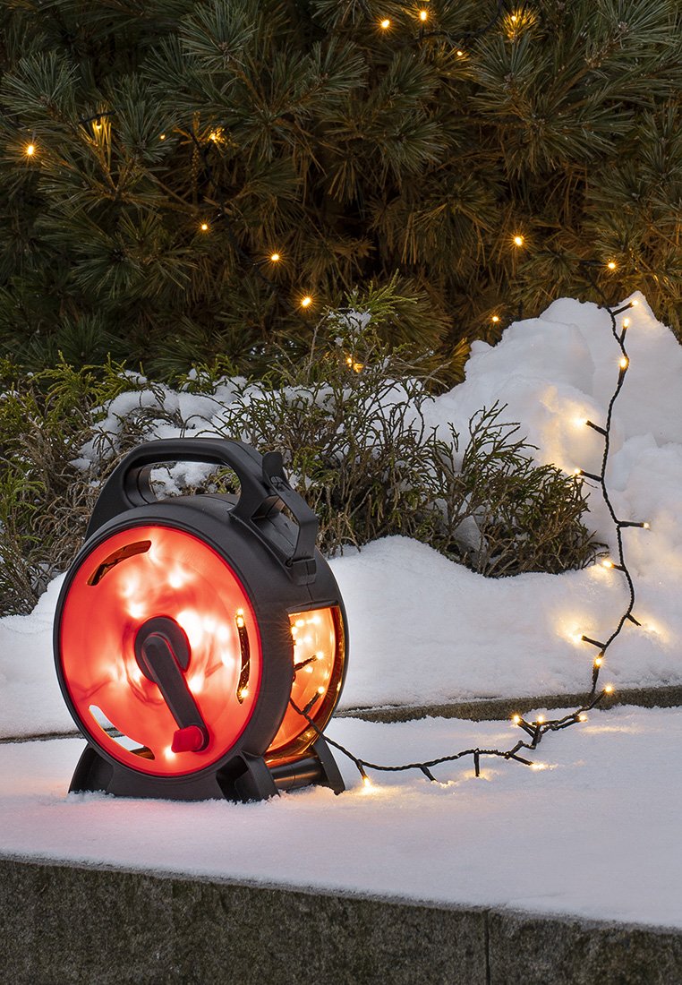 Carrete eléctrico portátil negro y rojo con luces expuestas en su interior, colocado sobre nieve blanca, con una cadena de luces enrollada a su lado.