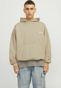 Béžová oversized mikina s přední kapsou, žebrovanými manžetami a lemem. Má malé bílé logo na hrudi. Nosená s lehkými denimovými cargo kalhotami.
