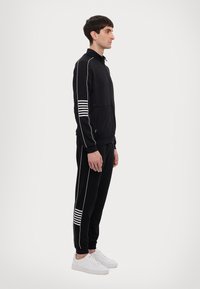 Completo sportivo nero con strisce bianche, composto da una giacca con zip e pantaloni abbinati. Il tessuto ha una consistenza liscia e un design aderente.