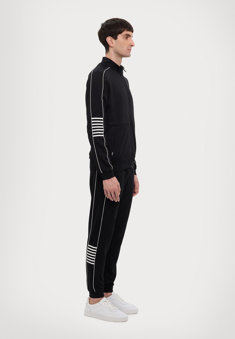 Completo sportivo nero con strisce bianche, composto da una giacca con zip e pantaloni abbinati. Il tessuto ha una consistenza liscia e un design aderente.