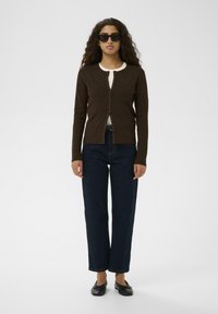 Ruda briaunota cardigan'ą ilgomis rankovėmis ir priekyje su mygtukais, derinamas su tamsiai mėlynos aukšto juosmens džinsais. Modelis vilki baltą marškinėlį po juo.