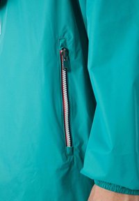 Giacca turchese con una tasca con cerniera caratterizzata da una zip bianca e rossa. Il tessuto appare liscio con una leggera lucentezza. I polsini sono elastici.