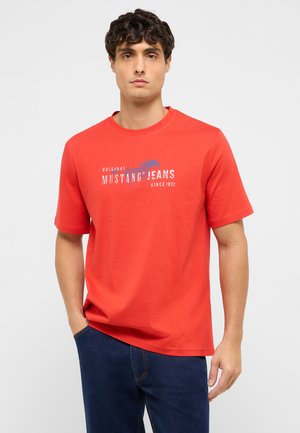 Mann trägt ein rotes Mustang Jeans T-Shirt mit weißem und blauem Logo-Schriftzug, kombiniert mit dunkelblauen Jeans, stehend vor einem einfachen Hintergrund.