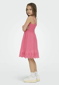 Růžové bavlněné šaty se smocked živůtkem, ramínky a krajkovým spodním lemem. Modelka je kombinuje s bílými kotníkovými ponožkami a hrubými bílými botami.