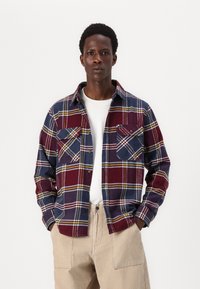 Rød og marineblå ternet flannel skjorte med gule accenter, to brystlommer og knaplukning foran, båret over en hvid T-shirt og beige kordfløjl bukser.