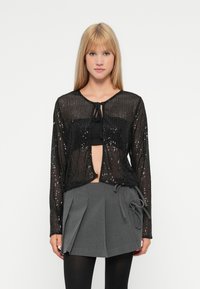 Cardigã preto com lantejoulas, com design cropped, mangas longas e fecho atado no pescoço, sobre uma blusa preta e uma saia plissada cinza.