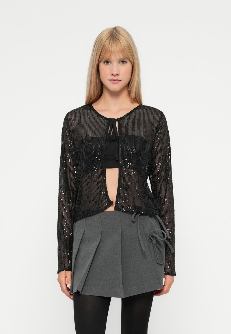 Cardigã preto com lantejoulas, com design cropped, mangas longas e fecho atado no pescoço, sobre uma blusa preta e uma saia plissada cinza.