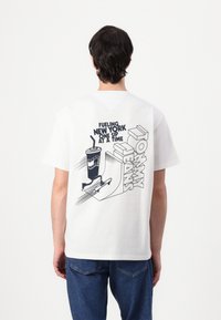 Hvid bomulds T-shirt med grafisk design på ryggen, der inkluderer tekst og illustrationer af en drink og et skateboard. Klassisk crewneck stil.