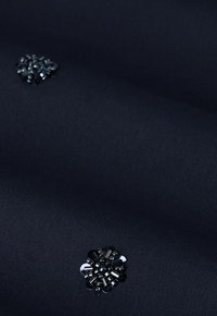 Marine stof met een gestructureerd oppervlak, versierd met een decoratieve zwarte bloemversiering van kralen en metalen elementen.