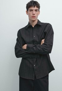 Ung mand med kort mørkt hår, der står foran en ensfarvet væg, iført en mørk skjorte med knapper og sorte bukser, med armene krydset.