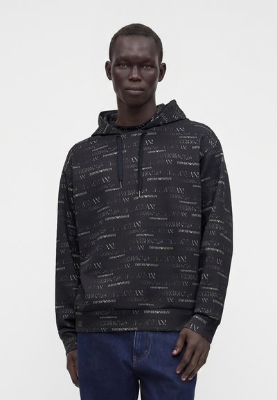 Άνδρας που φοράει μαύρη φούτερ Emporio Armani με επαναλαμβανόμενο κείμενο της μάρκας, συνδυασμένη με σκούρα μπλε τζιν, στέκεται μπροστά σε ένα απλό λευκό φόντο.