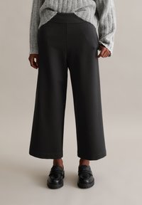 Pantalons larges noirs avec un waistband élastique, fabriqués dans un tissu lisse et extensible. Avec des mocassins noirs.