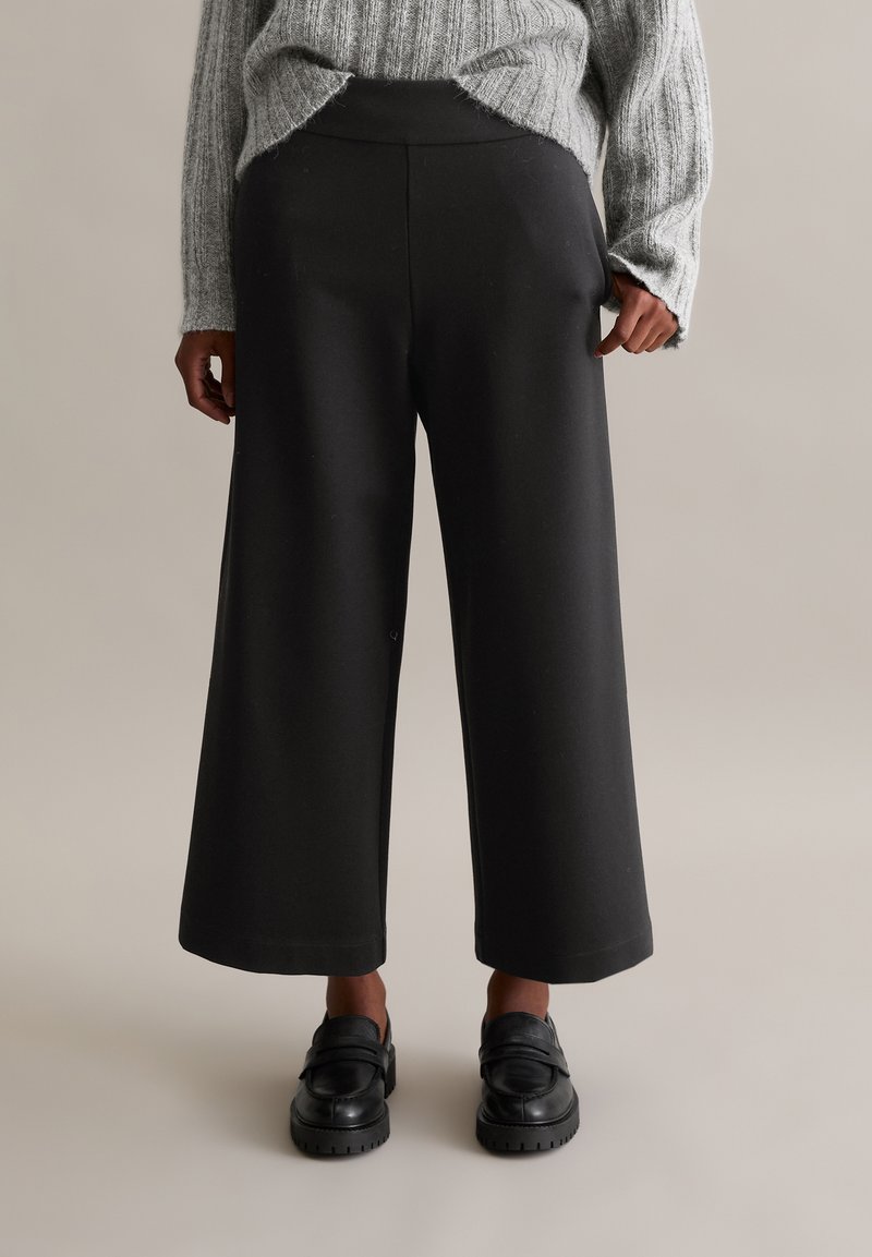Pantalons larges noirs avec un waistband élastique, fabriqués dans un tissu lisse et extensible. Avec des mocassins noirs.