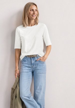 Kvinde med blondt hår iført en hvid skjorte og lyseblå jeans, der holder en grøn jakke, stående mod en ensfarvet lys væg.