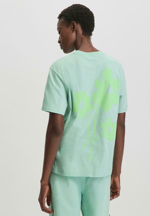 Minzgrünes Baumwoll-T-Shirt mit kurzen Ärmeln, das auf der Rückseite eine große, hellgrüne Blumengrafik zeigt. Weicher, lässiger Schnitt.