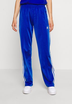 Vrouw draagt blauwe Adidas trainingsbroek met witte strepen langs de zijkanten en witte sneakers, staand tegen een eentonige achtergrond.