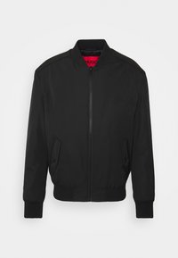 Giacca bomber nera con chiusura a zip, polsini e orlo a costine, tasche laterali angolate e etichetta interna rossa. Tessuto liscio e leggero.