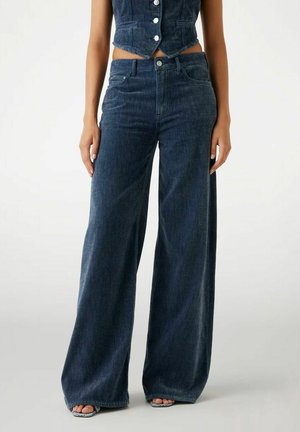 Kvinde iført højtaljede, vide mørkeblå jeans kombineret med en kort, knaplukket mørkeblå vest og åbne sandaler.