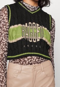Černý pletený zkrácený svetr se zelenými a béžovými pruhy s textem "RAGGED JEANS". Nosit na vzorovaném průhledném topu. Viditelné lehké kalhoty.