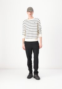 Suéter branco com riscas azul-marinho, punhos canelados, combinado com calças escuras e ténis pretos com padrão, complementado com um boné cinza.