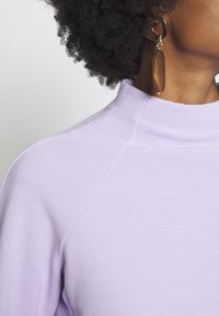 Lila ribbed Oberteil mit hohem Halskragen und strukturiertem Design, ergänzt durch einen großen runden goldenen Ohrring und einen Holzanhänger.